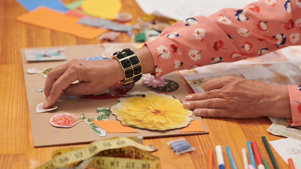 A female hand, with a pink floral long sleeve top, placing a cut out flower image on a sheet of card. There are various pencils and scrap paper scattered on the table.